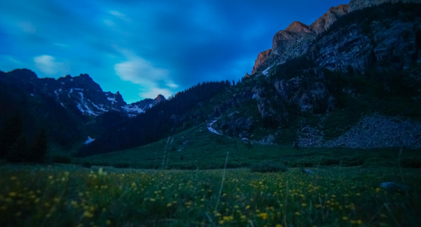 The sky is softly lit, illuminating a green meadow dotted with yellow wildflowers. There is a mountainous landscape surrounding the meadow.